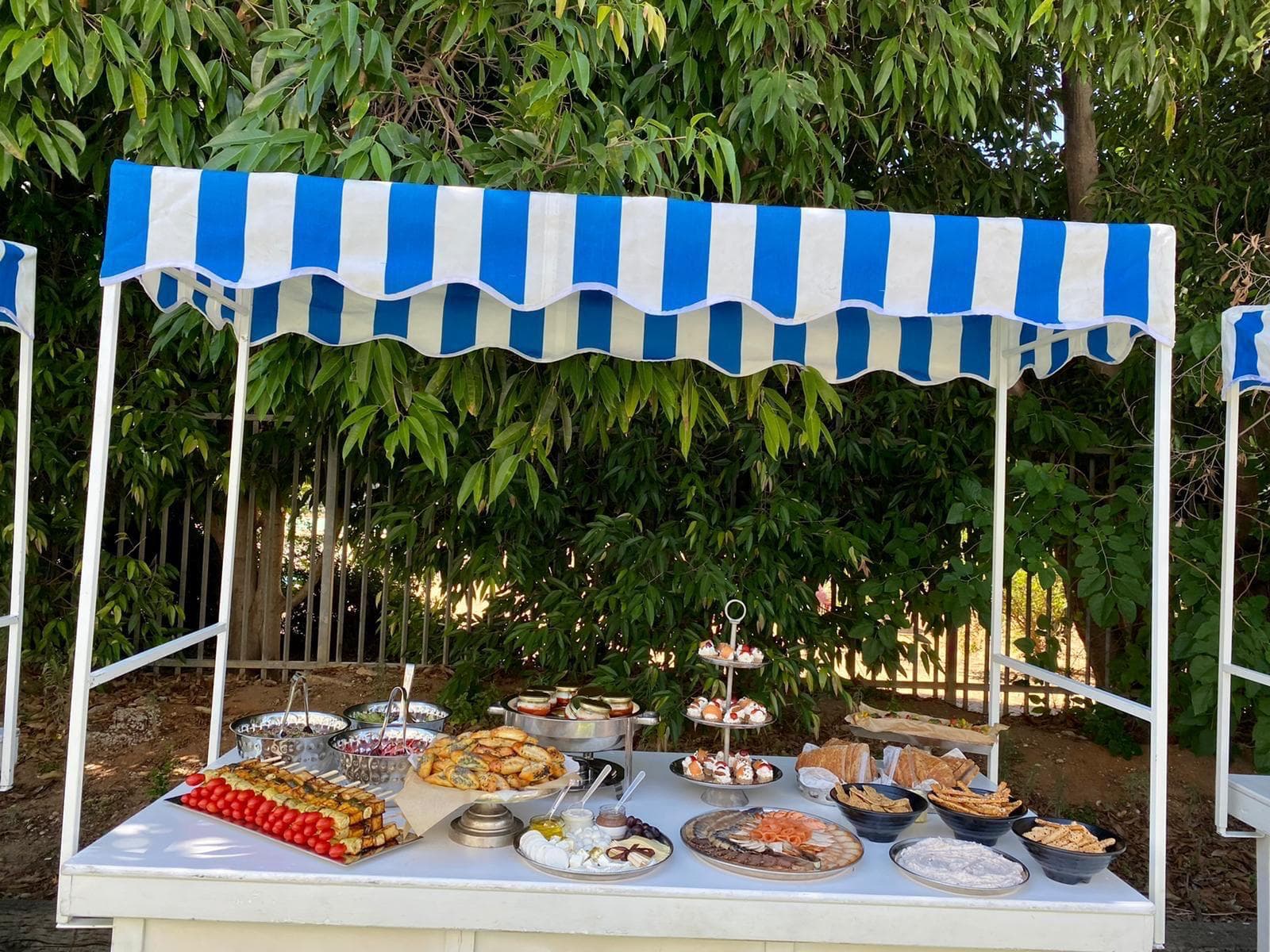 דוכן אוכל מעוצב לאירועים, עם מגוון מנות וקינוחים, תחת סככה עם פסים כחולים ולבנים.